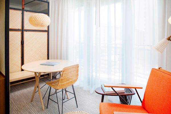 The image shows a bright room with an orange chair, a round table with a wicker chair, and a modern light fixture by a window with sheer curtains.