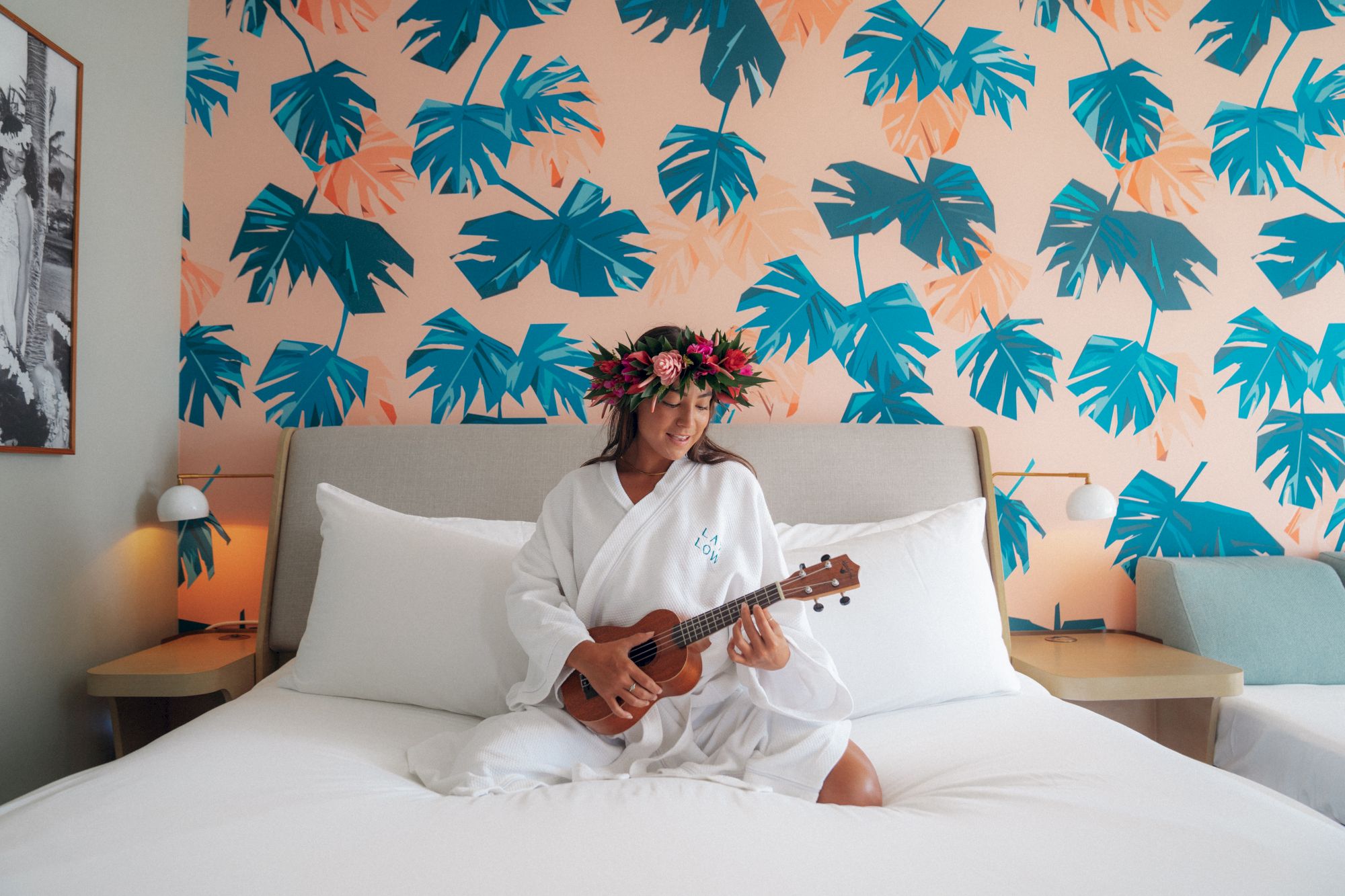 A person in a white robe sits on a bed playing a small ukulele in a tropical-themed bedroom with blue and peach palm leaf wallpaper, wearing a flower crown.