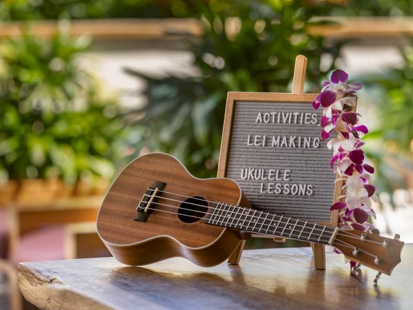 A ukulele rests on a table beside a sign with activities: "Lei Making" and "Ukulele Lessons," adorned with orchids.