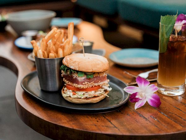 A juicy burger with fries and a drink on a plate, with a purple flower garnish beside it.