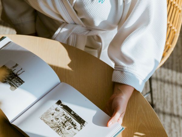 A person in a white robe is sitting at a table, holding an open book with black-and-white photographs, with sunlight casting shadows.