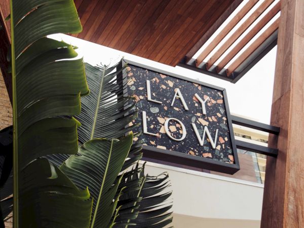 The image shows a sign reading "LAY LOW" with a terrazzo design, next to large green leaves and wooden architectural elements.