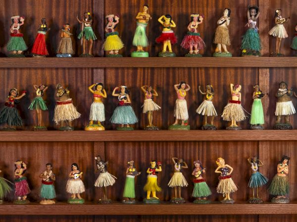 The image shows a collection of miniature hula dancer figurines in various poses, displayed on wooden shelves.