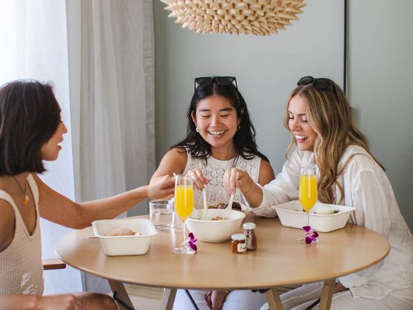 Three people sit at a round table enjoying drinks, surrounded by bright decor, and sharing a cheerful moment together.