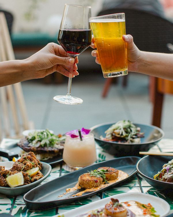 People toast with a glass of red wine and a beer over a table filled with various dishes and a cocktail.