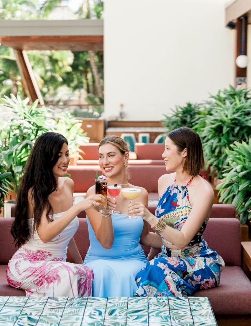 Three women sit on a couch outdoors, clinking wine glasses and chatting, with lush potted plants around them.