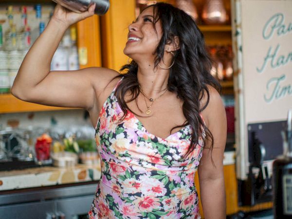 A woman in a floral dress is smiling while shaking a cocktail shaker in a bar setting.
