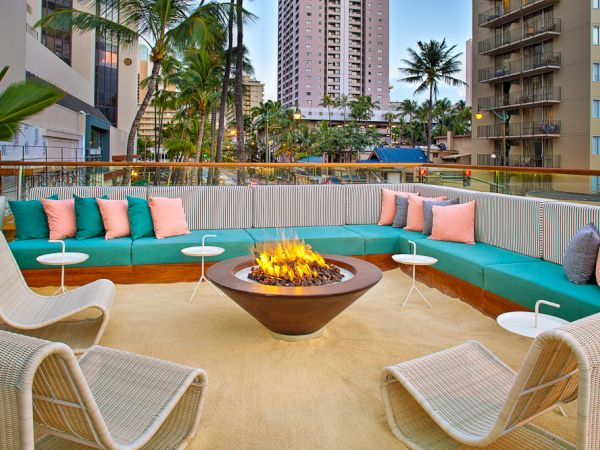 An outdoor lounge with a fire pit, surrounded by teal and pink cushioned seating, palm trees, and city buildings in the background.