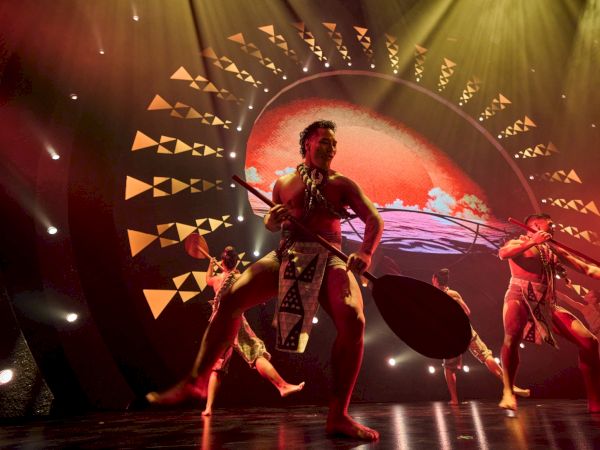 A group of performers in traditional attire dance on stage with paddles, under a large circular light and vibrant backdrop.