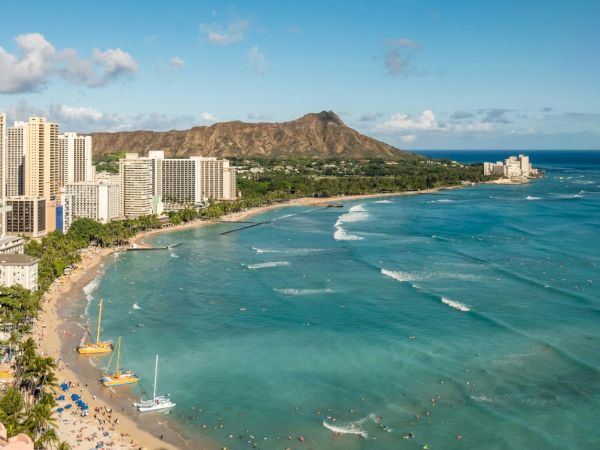 A beach with turquoise water, high-rise buildings, a lush mountain in the background, and boats along the shoreline.