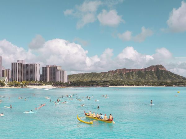 A vibrant beach scene with clear blue water, people kayaking and paddleboarding, skyscrapers, and a mountain in the background.