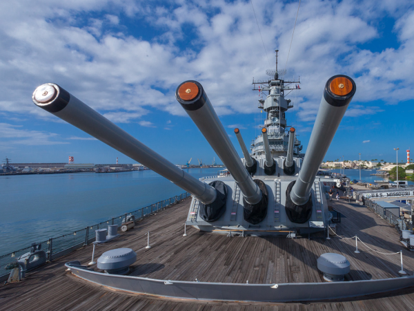 A battleship is shown with large artillery guns on its deck, docked in a harbor under a partly cloudy sky.