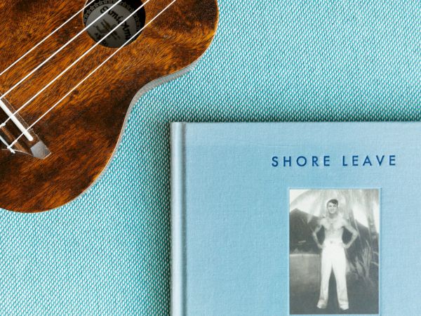 A ukulele lies next to the book "Shore Leave" by Ryan Mungia, featuring a vintage photograph on a blue textured background.