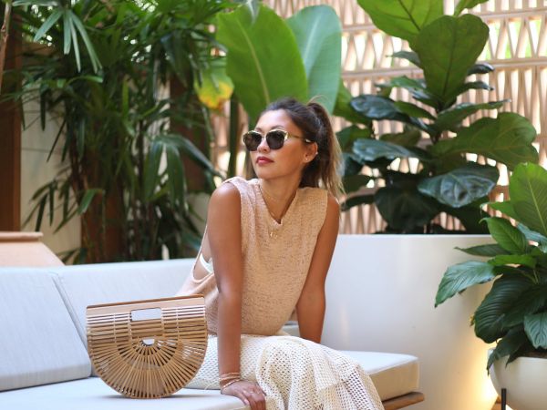 A person is sitting on an outdoor sofa surrounded by lush green plants, wearing sunglasses and casual attire, with a woven bag beside them.