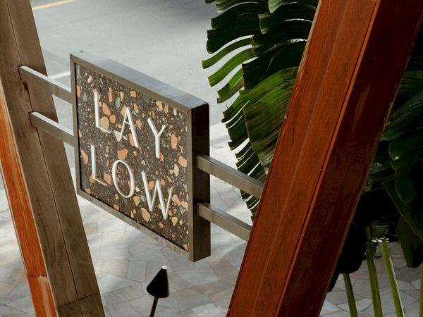 The image shows a sign with the words "LAY LOW" mounted on a structure, surrounded by wooden beams and tropical plants.