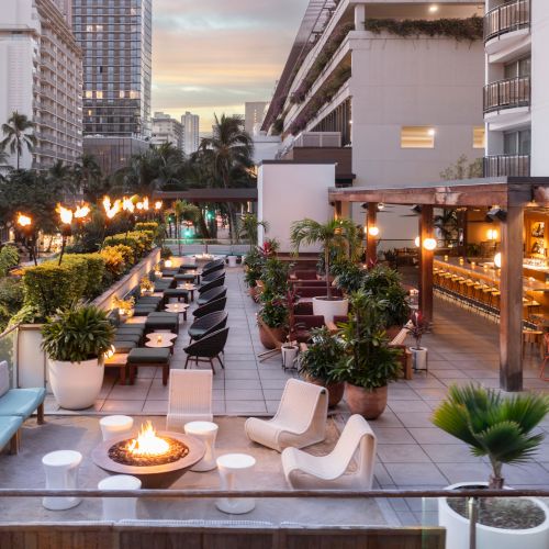 The image shows a rooftop terrace with stylish seating, a bar, a fire pit, and lush greenery, set against a cityscape backdrop at sunset.