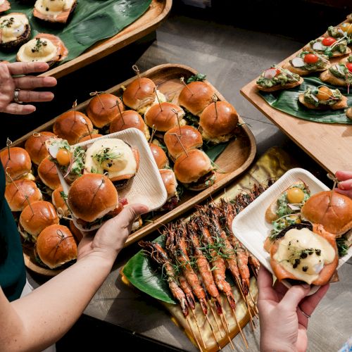 A spread of bite-sized sliders, grilled skewers, and mini bites on banana leaves and trays, with hands reaching in to share.
