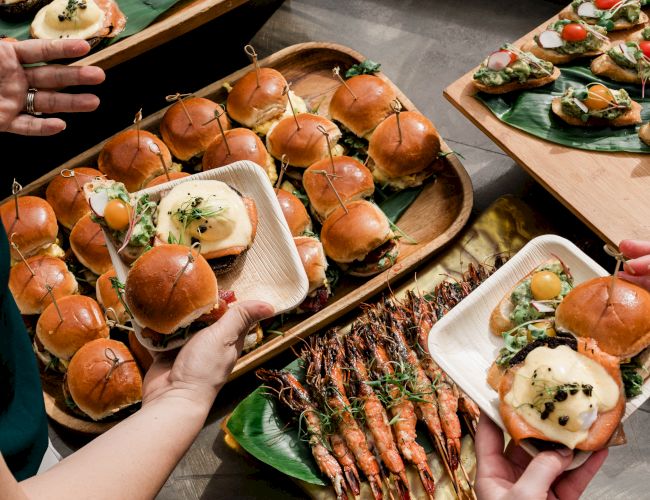 A spread of bite-sized sliders, grilled skewers, and mini bites on banana leaves and trays, with hands reaching in to share.
