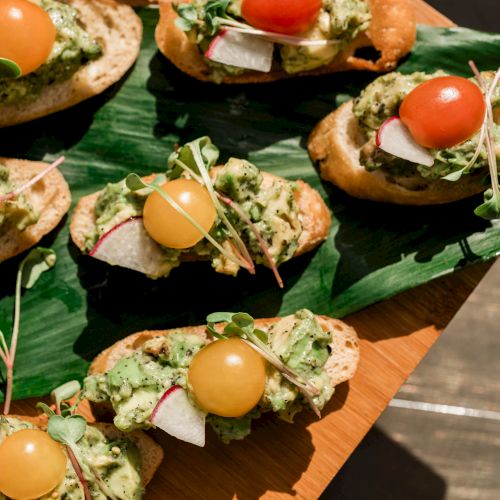 A platter of toasted bruschetta bites topped with creamy guacamole, cherry tomatoes, microgreens, and cheese, arranged on a wooden board.