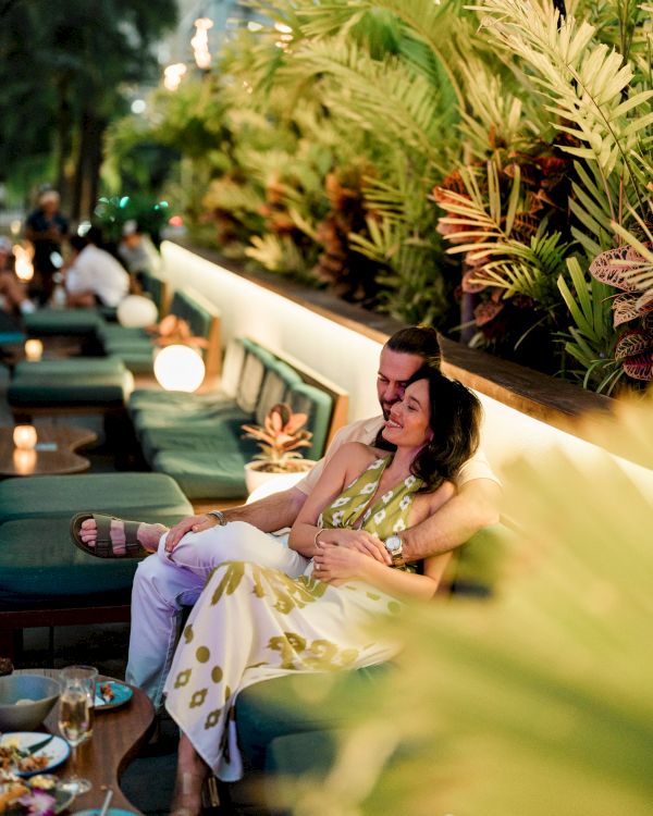 A couple relaxes at an upscale outdoor lounge, sipping drinks among tropical plants and soft ambient lighting.