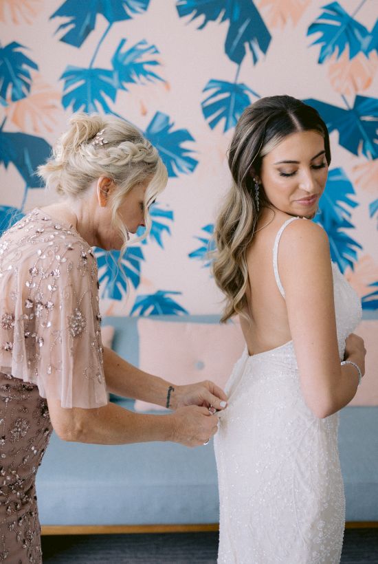 A woman in a light pink dress helps a bride with her wedding gown. They stand against a colorful wall with a leaf pattern.
