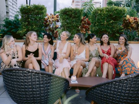 A group of nine people sitting on outdoor furniture, smiling and chatting, with greenery and decorative plants in the background.