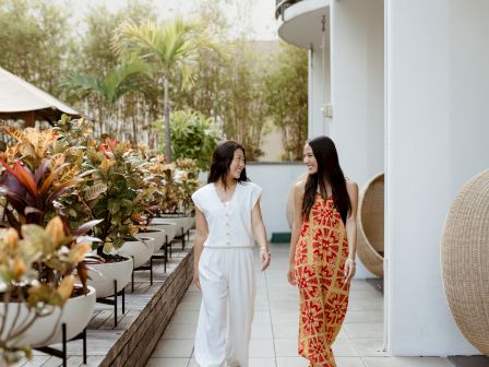 Two women stroll on a sunny outdoor patio, smiling at each other beside potted plants and palm trees.