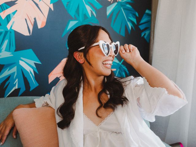 A woman in a white outfit posing on a couch with blue tropical wall art, wearing heart-shaped sunglasses and smiling.