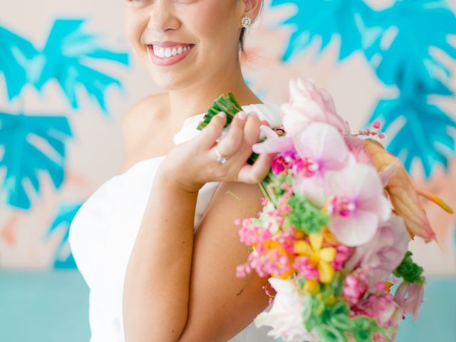 A smiling bride in a white dress holds a colorful bouquet against a blue tropical backdrop, posing with flowers adorning her shoulder.