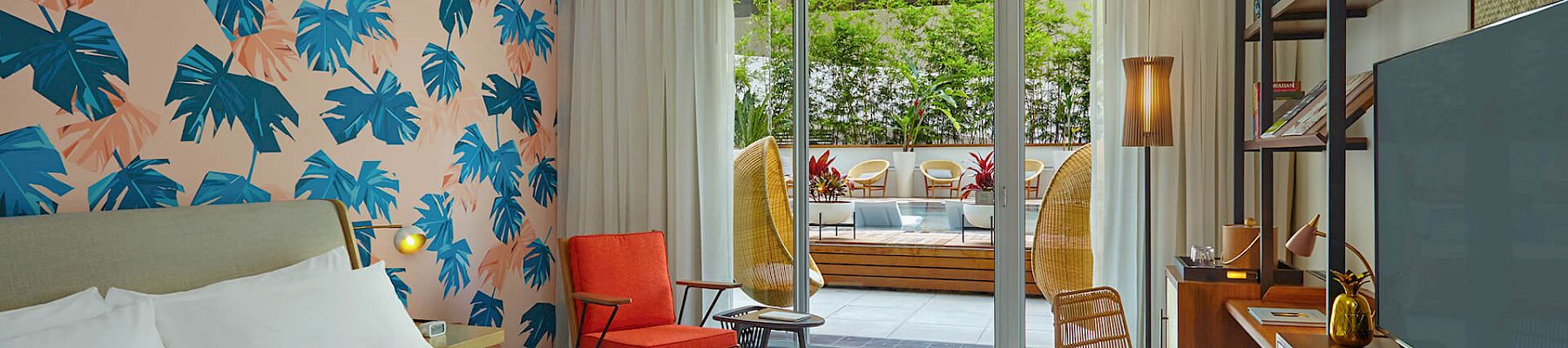 A modern hotel room with a tropical leaf wall design, a bed, TV, orange chair, and sliding doors opening to a balcony with seating.