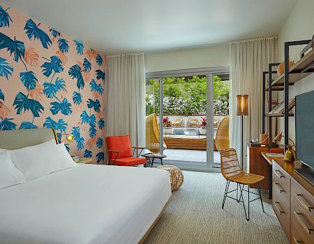 A modern hotel room with a tropical leaf wall design, a bed, TV, orange chair, and sliding doors opening to a balcony with seating.