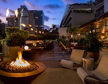 An outdoor lounge with a fire pit, cozy seating, and ambient lighting, set against an urban backdrop at dusk.