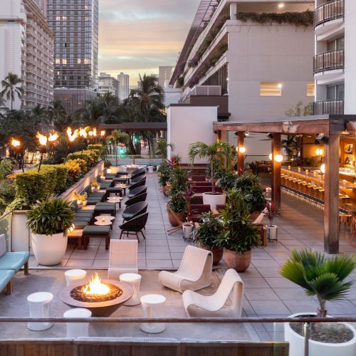 A stylish rooftop lounge with modern seating, string lights, potted plants, and a warm fire pit, overlooking city buildings at dusk.
