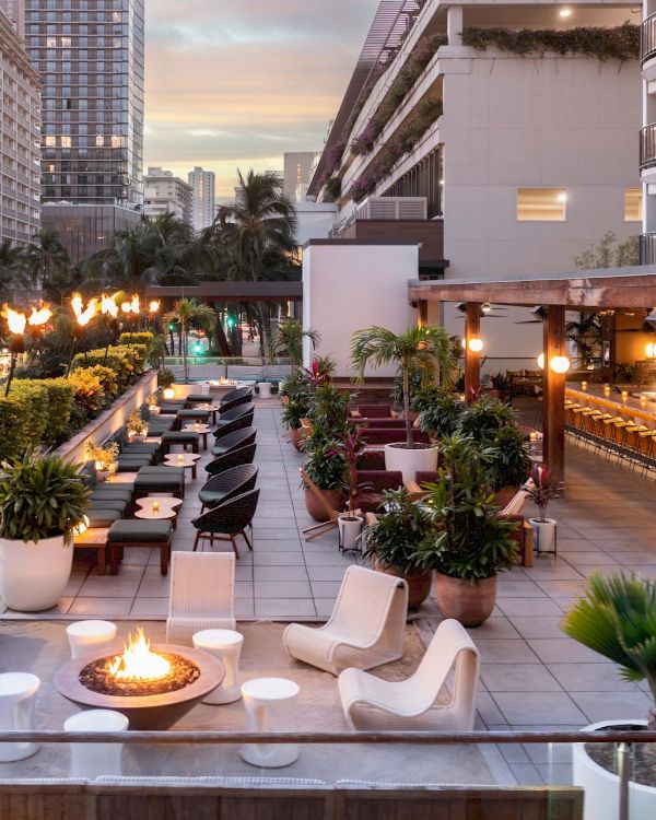 A stylish rooftop lounge with modern seating, string lights, potted plants, and a warm fire pit, overlooking city buildings at dusk.