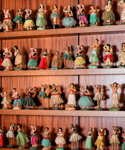 A wall of tiny, vintage dolls in colorful dresses lined up on wooden shelves, smiling faces and varied outfits.