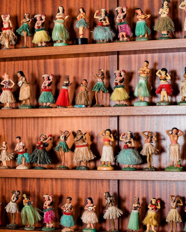A wall of tiny, vintage dolls in colorful dresses lined up on wooden shelves, smiling faces and varied outfits.