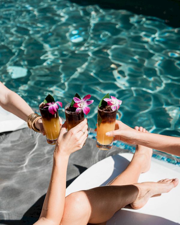 A sunny poolside scene with four people clinking tropical cocktails garnished with flowers, toes dipped in the water, bright and relaxed.