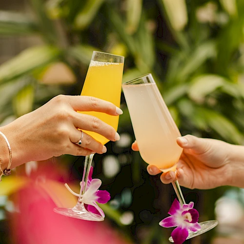 Two hands clink glasses with colorful drinks, adorned with flowers, against a lush, green background.
