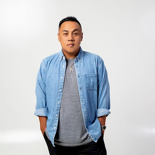 A person stands against a plain background, wearing a gray shirt and a blue denim button-up, hands in pockets.