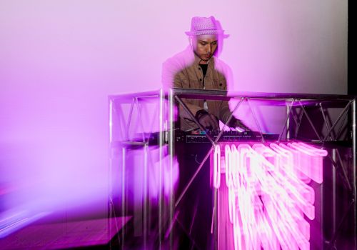 A DJ wearing a hat stands behind a glowing pink-purple neon-lit booth, spinning as vibrant light trails shoot out from the equipment.