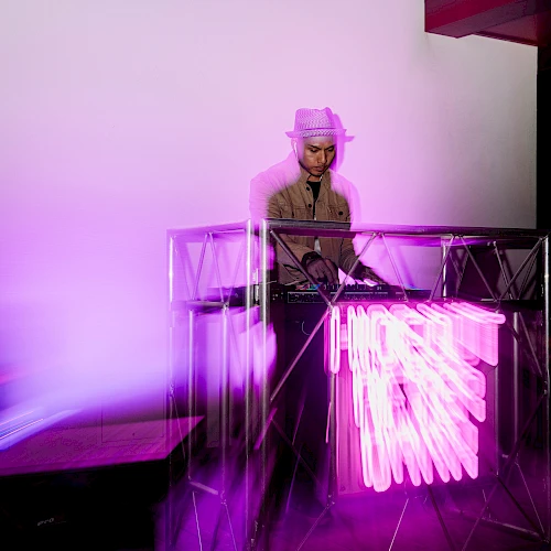 A DJ wearing a hat stands behind a glowing pink-purple neon-lit booth, spinning as vibrant light trails shoot out from the equipment.