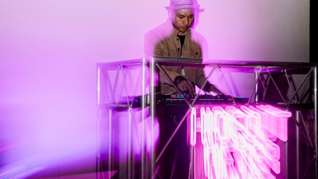 A DJ wearing a hat stands behind a glowing pink-purple neon-lit booth, spinning as vibrant light trails shoot out from the equipment.