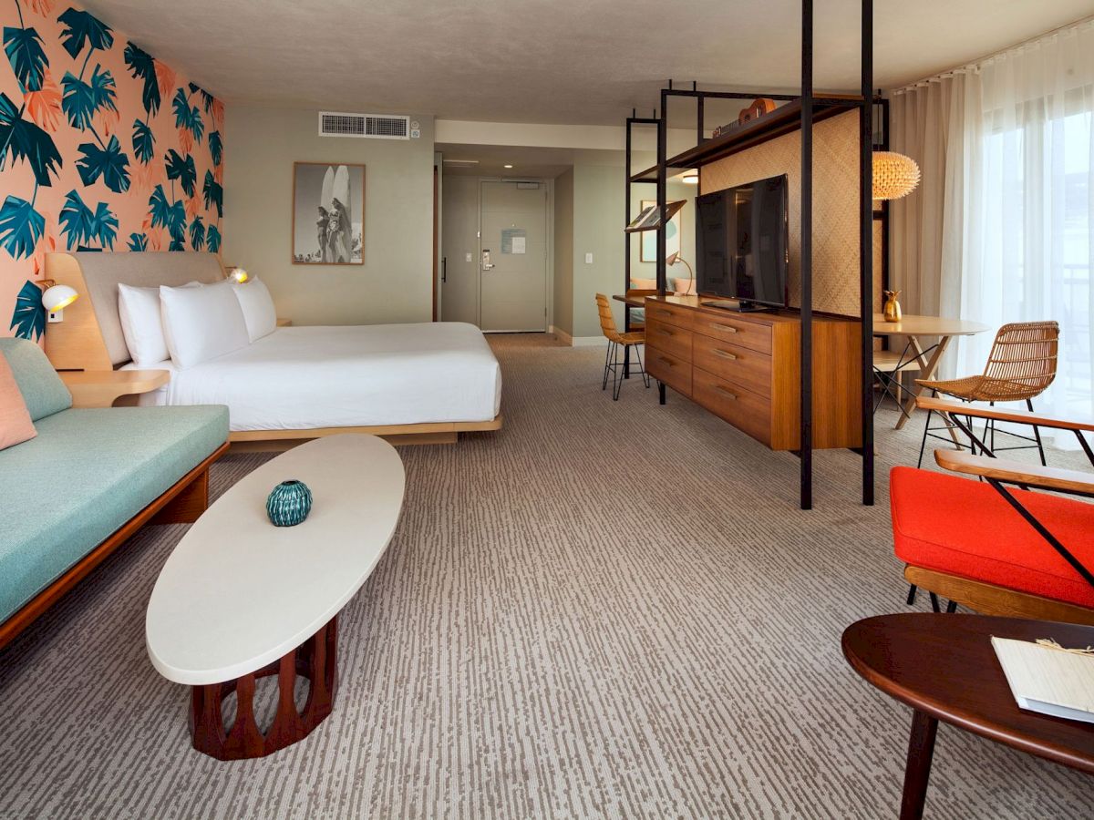 A spacious hotel room with colorful decor, featuring a bed, sofa, table, desk, and TV, with large windows and a patterned wall.