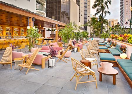 Open-air patio with colorful furniture, wooden chairs, and lush plants. Bar area with stools on the left, surrounded by tall buildings.