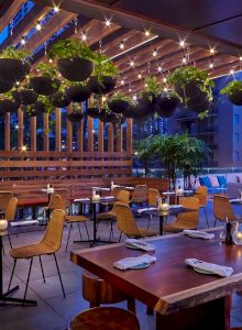 An outdoor restaurant patio with wooden tables, chairs, hanging plants, and string lights under a wooden pergola, creating a cozy atmosphere.
