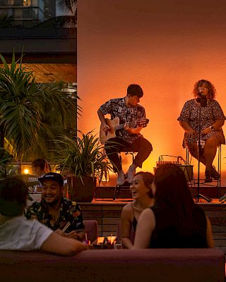 An outdoor setting with musicians performing for an audience at tables, surrounded by plants and warm lighting in the evening.