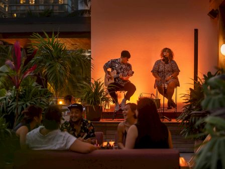 An outdoor setting with musicians performing for an audience at tables, surrounded by plants and warm lighting in the evening.
