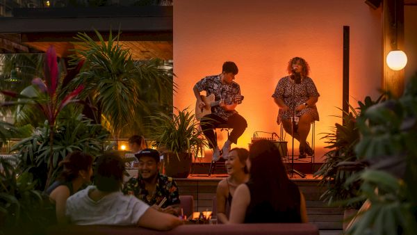 A cozy rooftop scene at dusk with a small acoustic duo performing on a warm-lit stage while a group lounges on a couch, watching and chatting.