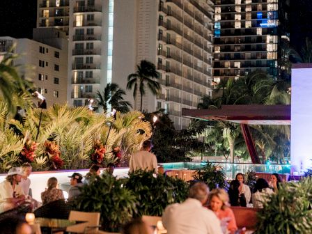 Nightlife rooftop scene with people dining, palm trees, city buildings lit in the background, warm lanterns glow, relaxed social vibe.