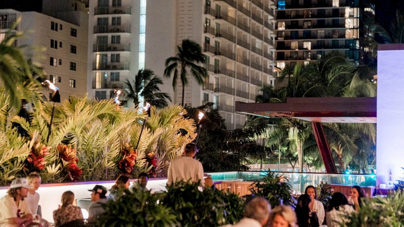 Nightlife rooftop scene with people dining, palm trees, city buildings lit in the background, warm lanterns glow, relaxed social vibe.
